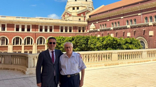 Two men in formal suits stand side by side in front of a large, modern structure