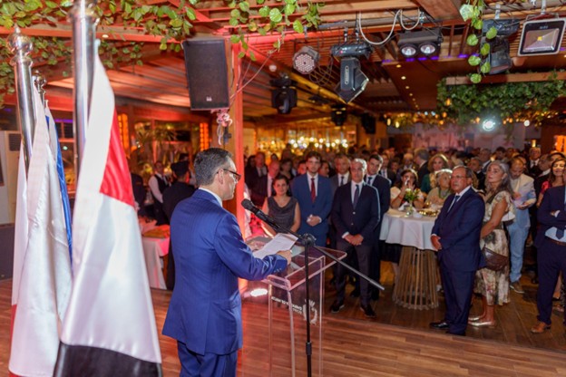 A man in a suit stands at a podium with a microphone, surrounded by people indoors.