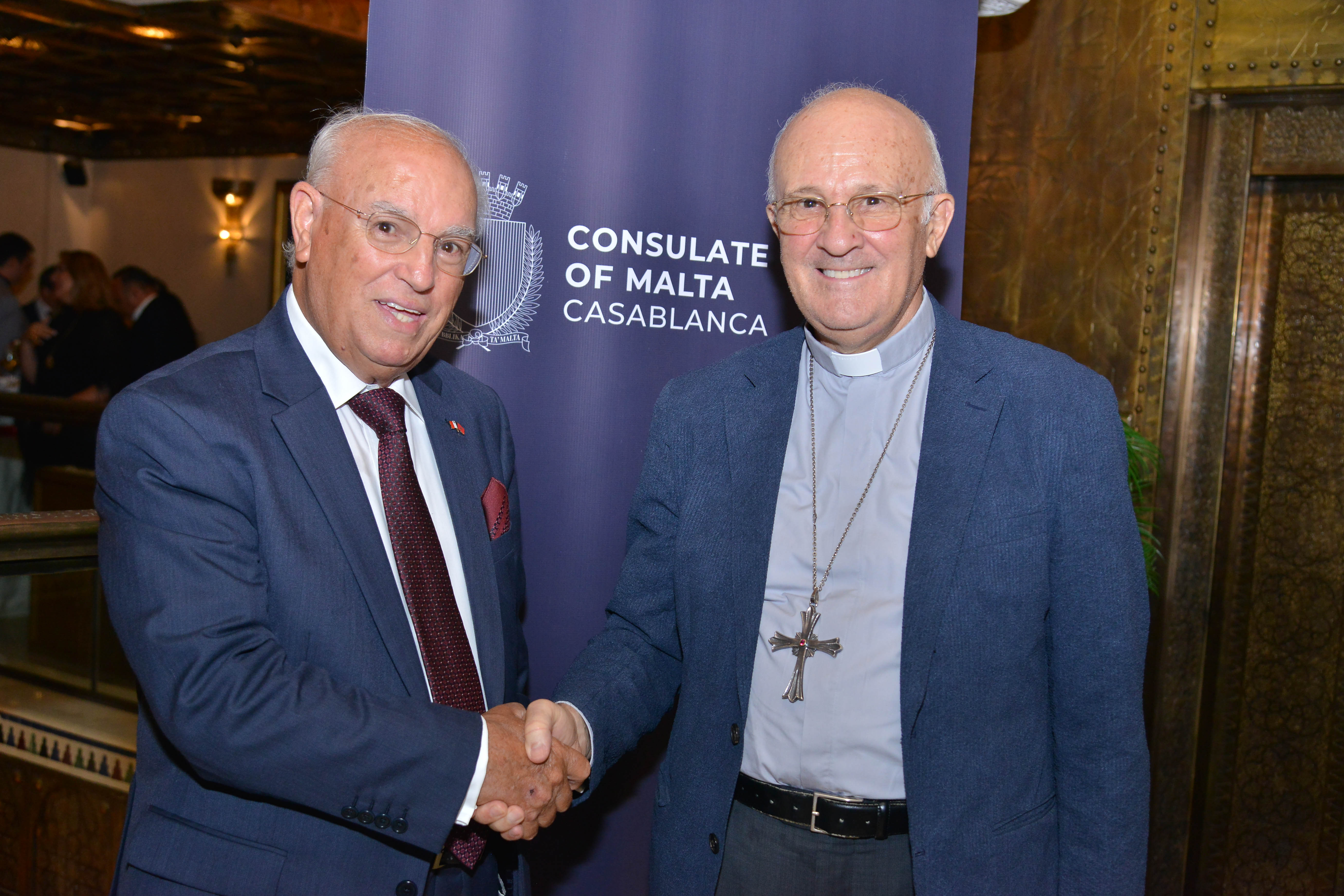 H.E. Paul Mifsud shaking hands with the Papal Nuncio Mons Alfred Xuereb with pull-up poster of Consulate in background.