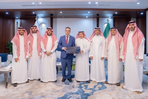 A group of eight men stands in a formal room. Seven wear traditional Saudi attire while one is in a blue suit. The mood is formal and official.