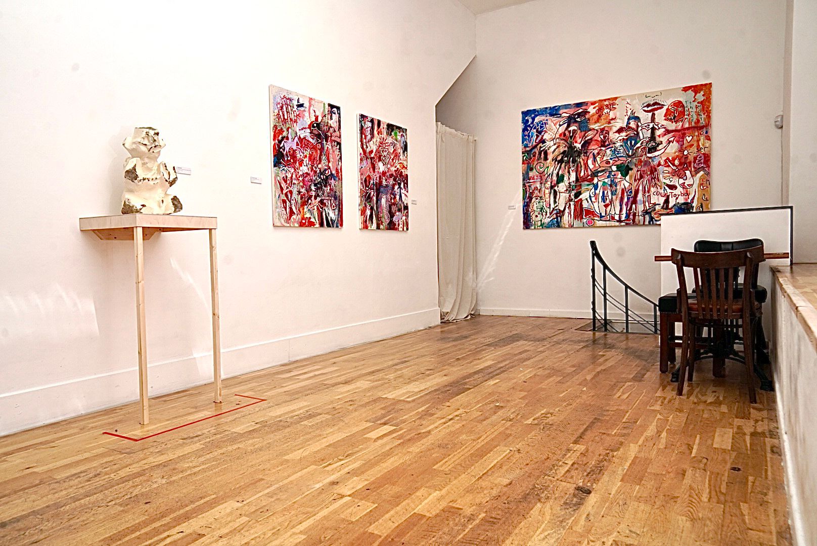 A gallery space with a small abstract white statue on a table to the left, two small abstract multicoloured paintings on the left wall and a larger abstract muticoloured painting on the back wall. A table and two chairs stand in front of the larger painting.