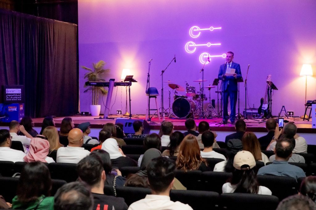 Photograph shows a live stage presentation in a purple-lit venue, with a speaker at a lectern addressing a seated audience. Stage includes drum kit, keyboards, guitars, microphones, potted plant, floor lamps, and three glowing circular neon symbols on back wall