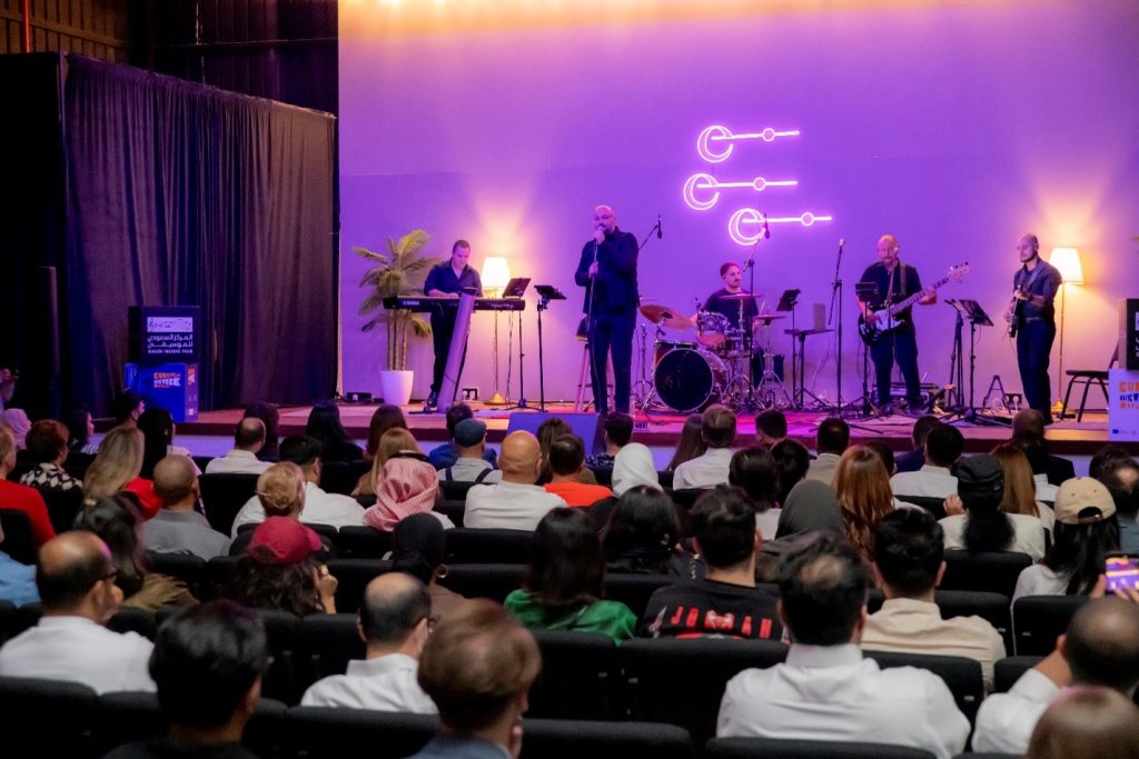 Photograph of a live band performing onstage before a seated audience in an indoor venue. Purple and amber lighting, neon-style musical logo, vocalist, keyboardist, drummer, guitarists, microphones, and instruments define concert setting.