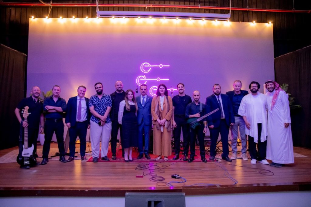 Photograph of a diverse group of musicians and performers posing on a theater stage after a concert or cultural event. Purple and warm stage lighting, projected musical symbols, instruments, microphones, and varied formal, casual, and traditional attire emphasize a collaborative international performance.